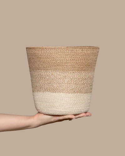 Eine Hand hält den Pflanzenkorb aus Jute (Lapis | 22 cm Ø), mit beigen und cremefarbenen Streifen auf taupefarbenem Hintergrund. Dieser handgefertigte Korb bietet ein strukturiertes Design, das perfekt dazu geeignet ist, jedem Raum eine organische Note zu verleihen.