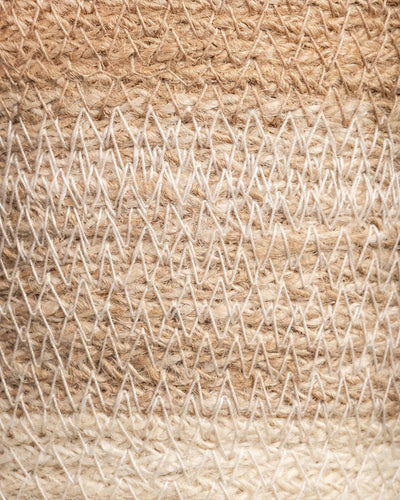 Nahaufnahme eines gewebten Textils mit Zickzackmuster in Beige- und Cremetönen, das an die Textur des Pflanzenkorbs aus Jute (Lapis | 22 cm Ø) erinnert. Die groben Fasern erzeugen ein komplexes Aussehen, wobei helle und dunkle Farbtöne Tiefe verleihen.