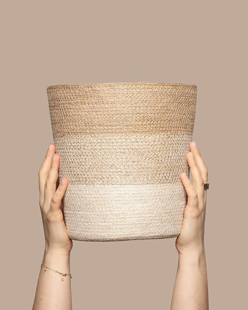 Zwei Hände halten einen Pflanzenkorb aus Jute (Lapis | 25 cm Ø) mit horizontalen hell- und dunkelbeigen Streifen. Der schlichte taupefarbene Hintergrund betont die natürliche Jutestruktur des Korbes.