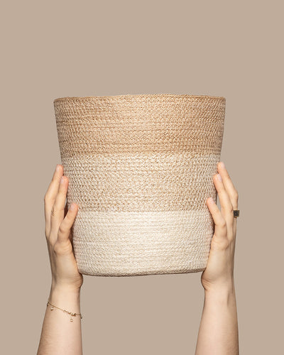 Zwei Hände halten einen Pflanzenkorb aus Jute (Lapis | 25 cm Ø) mit horizontalen hell- und dunkelbeigen Streifen. Der schlichte taupefarbene Hintergrund betont die natürliche Jutestruktur des Korbes.