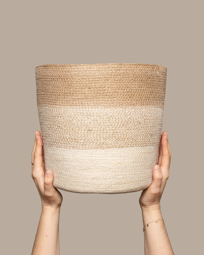 Zwei Hände halten einen Pflanzkorb aus Jute (Lapis | 30 cm Ø) mit einem Farbverlauf von Beige nach Hellcreme vor einem schlichten Taupe-Hintergrund.