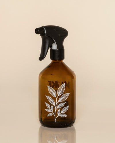 Der Pflanzensprüher aus Amber Glas mit schwarzem Sprühkopf und weißem botanischem Blattmotiv ist das stilvolle Zubehör zur Pflanzenpflege - präsentiert vor schlicht beigem Hintergrund.