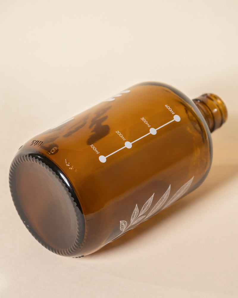 Eine braune Glasflasche Pflanzensprüher liegt auf der Seite und zeigt eine weiße Messskala mit beschrifteten Linien und Punkten sowie zarte botanische Illustrationen in der Nähe des Bodens. Der leere Sprüher hat eine große Öffnung.