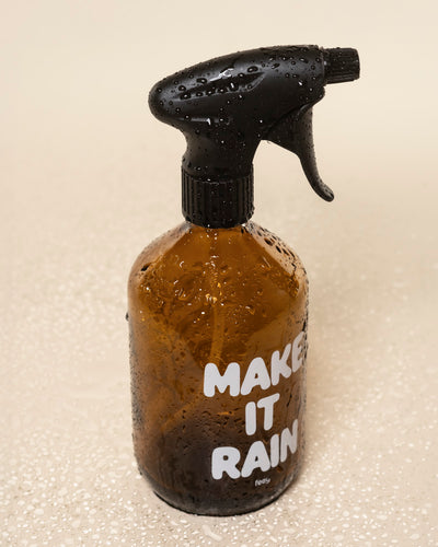 Ein brauner Pflanzensprüher mit schwarzer Düse und weißem "MAKE IT RAIN"-Schriftzug sitzt auf einer nassen, hellen Fläche. Seine Wassertropfen unterstreichen den perfekten Einsatz für die Pflege Ihrer Lieblingspflanzen.