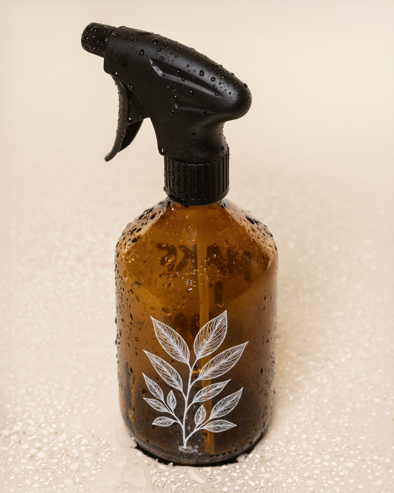 Ein Pflanzensprüher aus braunem Glas mit schwarzem Sprühkopf und weißem botanischen Pflanzen-Motiv steht, von Wassertropfen bedeckt, auf einer nassen Fläche.