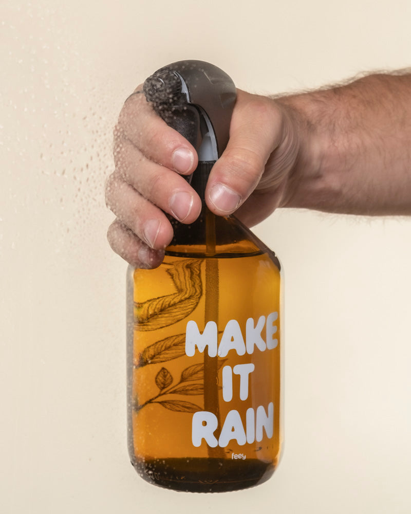 Eine Hand hält einen braunen Pflanzensprüher mit der Aufschrift "MAKE IT RAIN", Wassertropfen auf der Flasche und beschlagenes Glas auf der linken Seite.
