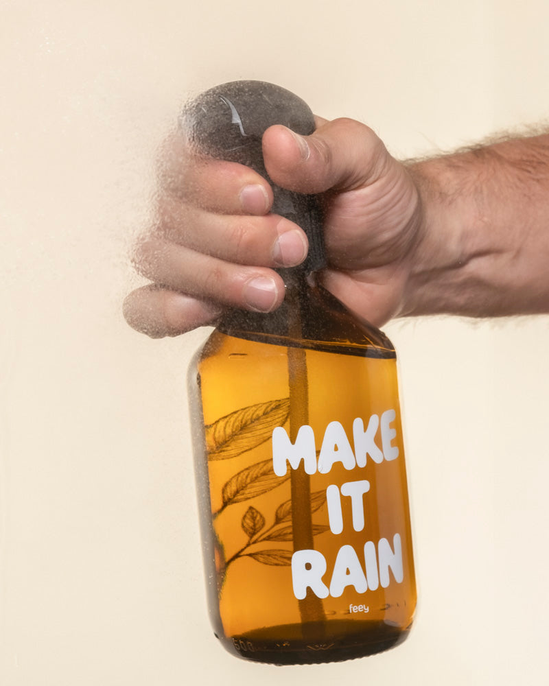 Eine Hand besprüht eine Pflanzensprher-Flasche mit der Aufschrift "MAKE IT RAIN" mit gelber Flüssigkeit; aus der gläsernen Düse tritt Nebel aus, ideal zum Gießen Ihrer Pflanzen.