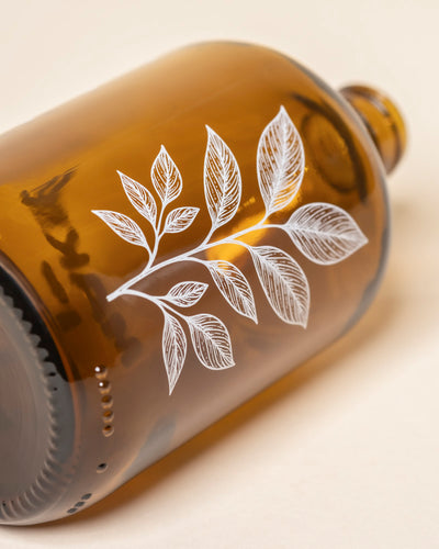 Eine braune Glasflasche namens Pflanzensprüher liegt seitlich und zeigt eine weiße Zeichnung eines Blattzweigs - ideal als dekorativer Pflanzensprüher für deine Pflanzen.