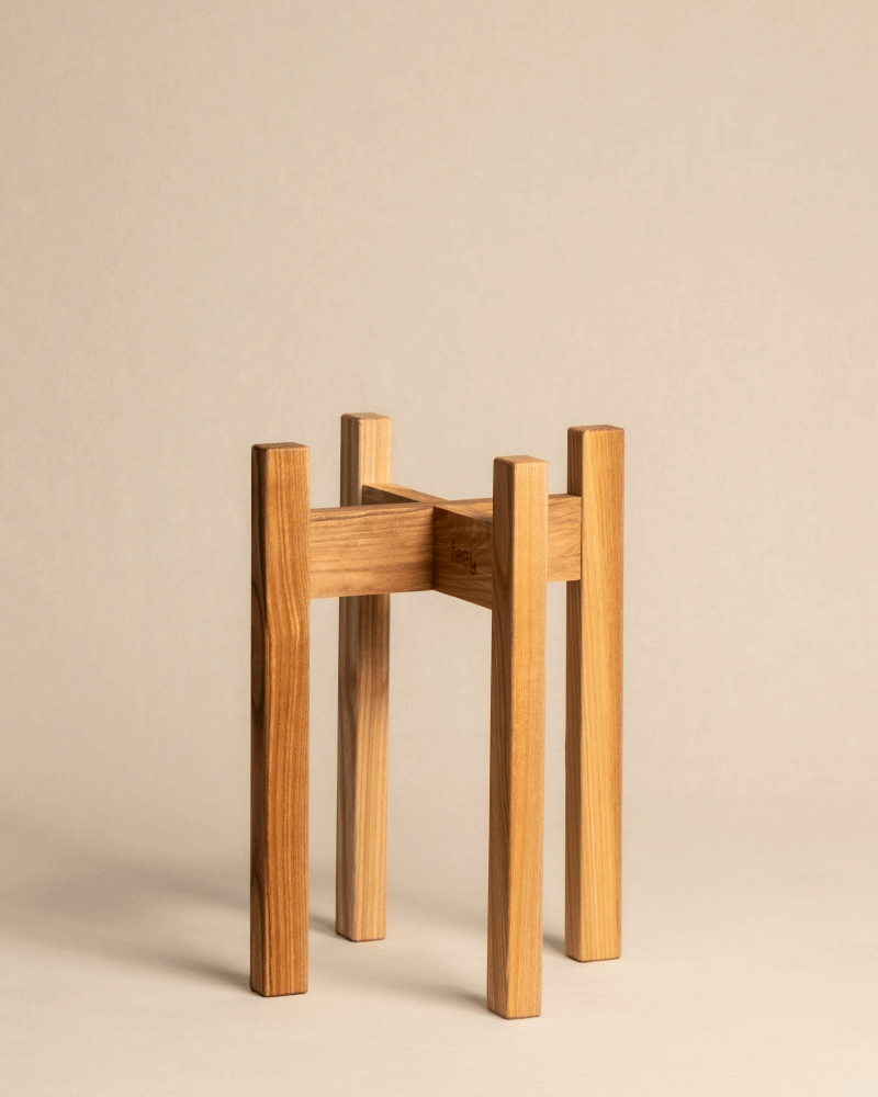 Ein Pflanzenständer aus der Stelion-Reihe mit minimalistischem Design und hohen, quadratischen Beinen aus Eschenholz vor einem beigen Hintergrund. Die natürliche Holzmaserung unterstreicht seinen rustikalen Charme und macht ihn zu einer idealen und stilvollen Wahl.