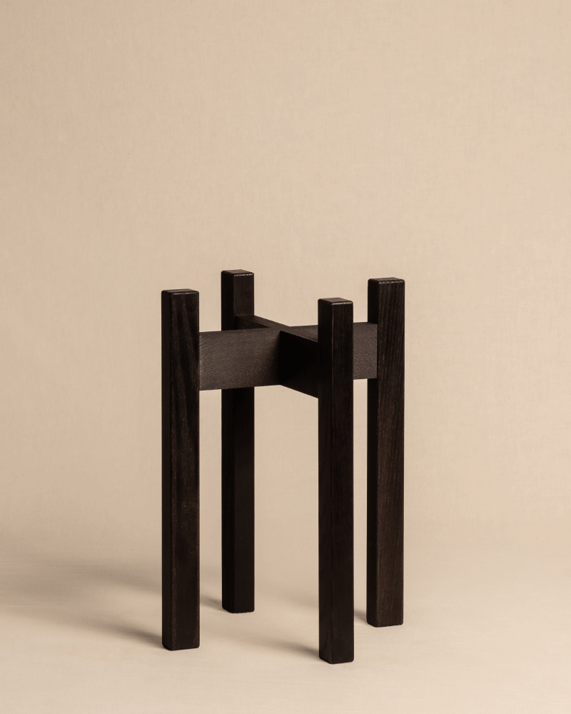 Das Produkt, Pflanzenständer (Stelion | 16 cm Ø), präsentiert ein zeitgenössisches Design mit minimalistischer Ästhetik. Es verfügt über vier hohe Eschenholzbeine und einen schlanken Holzsockel, alles vor einem neutralen beigen Hintergrund.