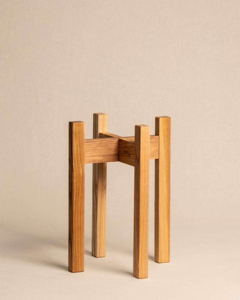 Ein minimalistischer Pflanzenständer aus Eschenholz, der ein schlichtes, modernes Design mit vier vertikalen Beinen und einer rechteckigen Stützstruktur aufweist. Das hellbraune Holz hat eine glatte Oberfläche und passt daher ideal zu den Töpfen 16 cm auf ihrem schlichten beigen Hintergrund.