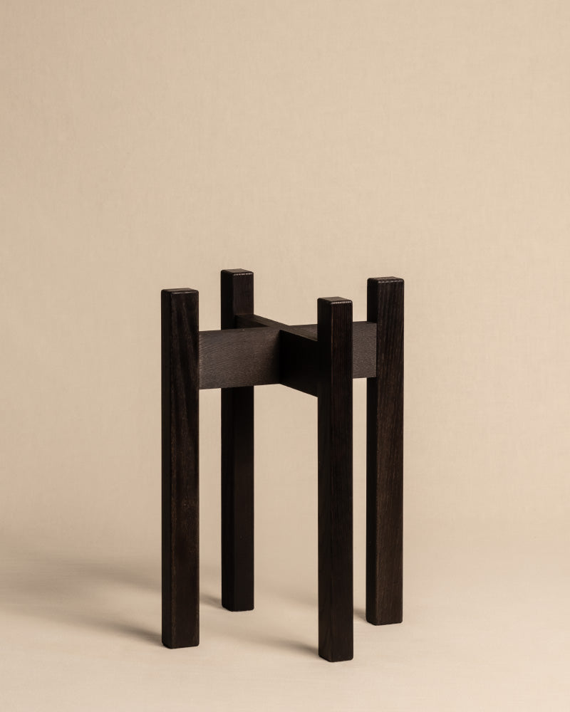 Ein minimalistischer Pflanzenständer (Stelion | 16 cm Ø) aus dunklem Eschenholz verfügt über vier hohe, rechteckige Beine und eine schlichte, flache Oberseite. Mit seinem geometrischen, modernen Design ergänzt dieses Stück jeden Raum als schicke Ergänzung vor einem neutralen beigen Hintergrund.