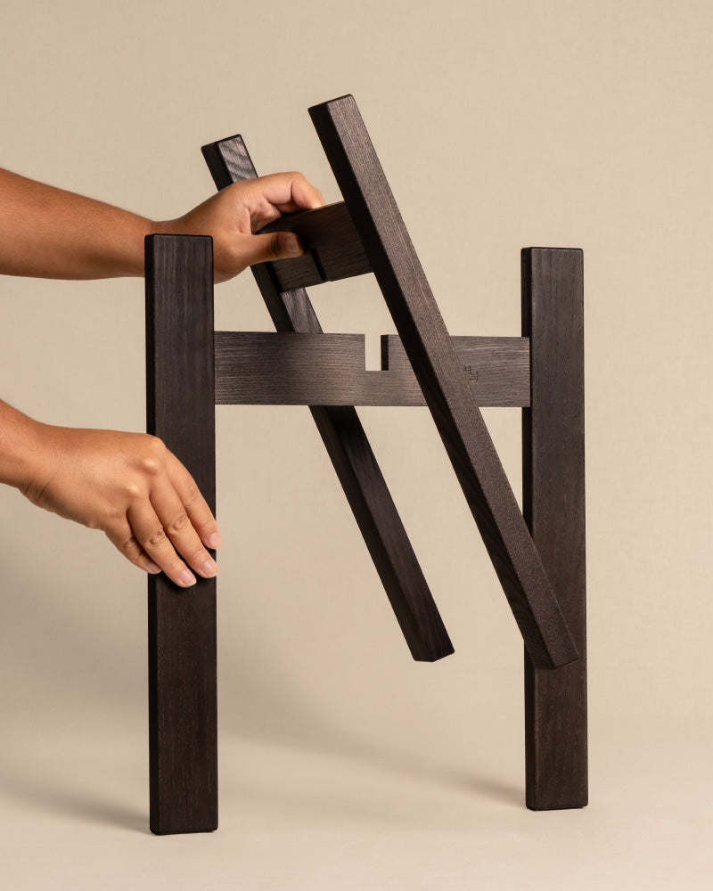 Hände montieren einen kleinen, dunklen Stelion Eschenholz-Rahmen vor einem beigen Hintergrund. Der Rahmen besteht aus ineinandergreifenden rechteckigen Teilen, die eine geometrische Struktur bilden, die sich perfekt als Stütze für Töpfe mit einer Größe von 16 cm eignet oder als eleganter Pflanzenständer (Stelion | 16 cm Ø) dient.