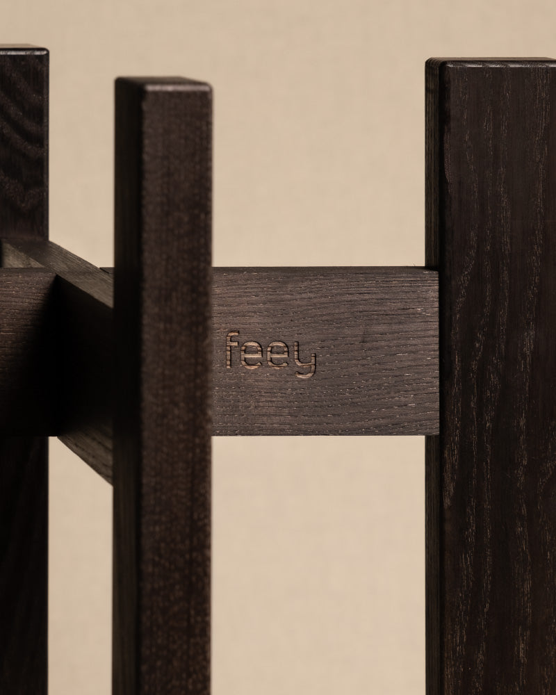 Nahaufnahme einer Holzkonstruktion aus Eschenholz mit dunkler Oberfläche, in deren einen der vertikalen Balken das Wort „feey“ eingraviert ist. Der Hintergrund ist in einem neutralen Beige gehalten, wodurch die Textur und die Verarbeitung hervorgehoben werden und an einen eleganten Pflanzenständer (Stelion | 16 cm Ø) erinnert.