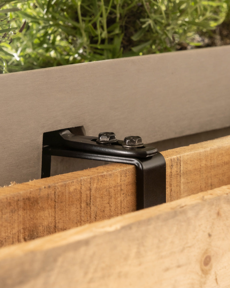 Nahaufnahme eines Metallbügels mit Schrauben zur Befestigung des Pflanzkastens für Geländer (Brügge | 48 cm) an Holzbrettern als Teil eines Hängesystems Balkon, mit Grünpflanzen im Hintergrund.