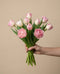 Eine Hand hält einen Strauß Tulpen "Liebesgedicht" - rosafarbene und weiße Tulpen mit grünen Stielen und Blättern - vor einem einfarbig beigen Hintergrund.