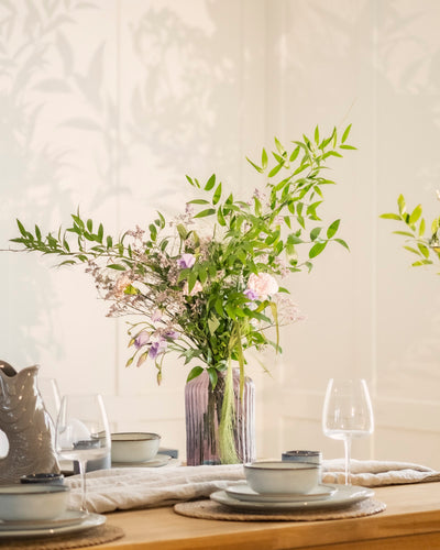 Eine mit grünen Blättern und pastellfarbenen Blumen gefüllte Purpurpoesie-Vase steht auf einem mit Tellern, Schüsseln, Gläsern und Tischsets gedeckten Esstisch in weichem Licht. Ihre Schatten werfen Anmut und Poesie auf die Wand dahinter.