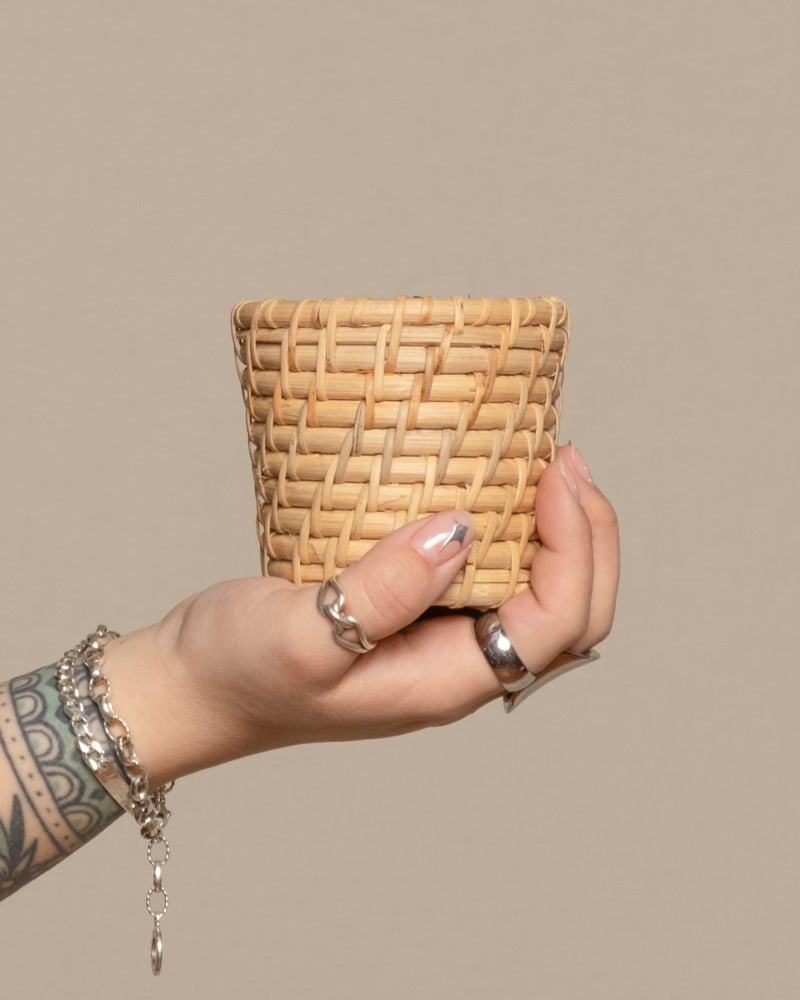 Eine tätowierte Hand mit silbernen Ringen und Armbändern hält einen kleinen Rattan-Babypflanzen-Korb „Calen“ vor einem schlichten beigen Hintergrund.