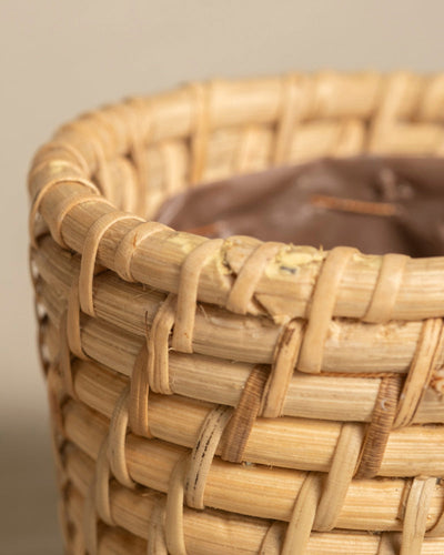 Nahaufnahme des Rattan-Babypflanzen-Korbs „Calen“, handgefertigt aus natürlichem, hellem Rattan mit strukturiertem Webmuster, das die Handwerkskunst hervorhebt und mit einem braunen Innenfutter ausgestattet ist.