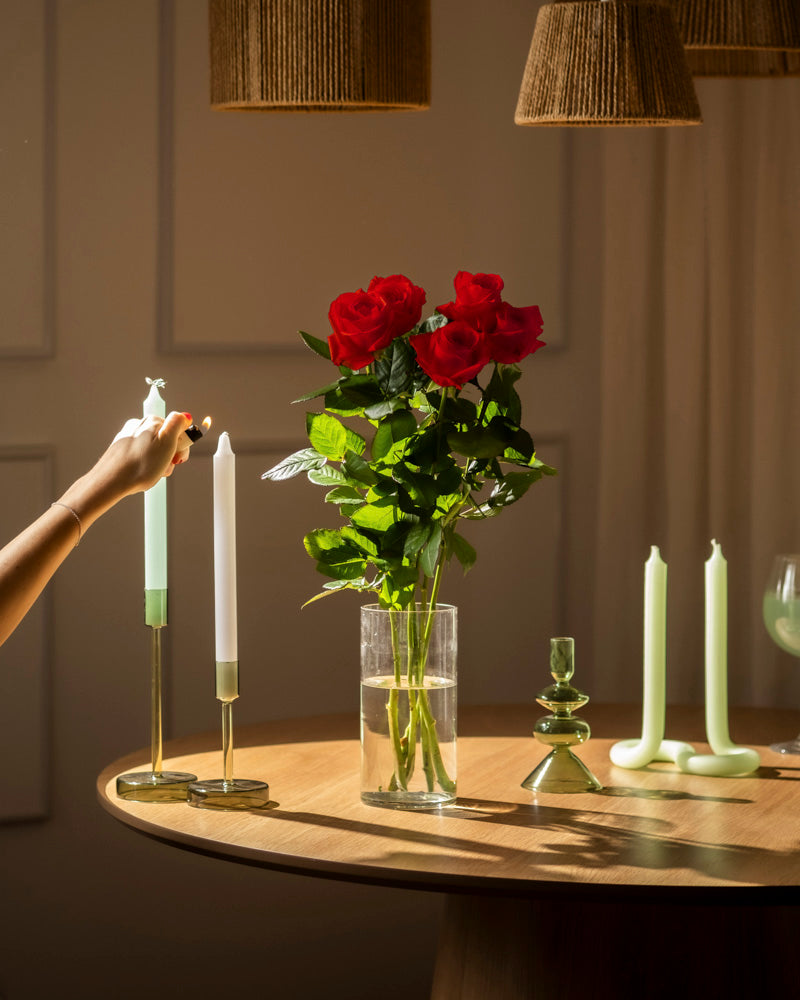 Eine Hand zündet eine weiße Kerze auf einem Holztisch an, der mit Roten Rosen (Inventar), grünen Kerzenhaltern und anderen Kerzen dekoriert ist, die alle sanft in warmem Licht leuchten.