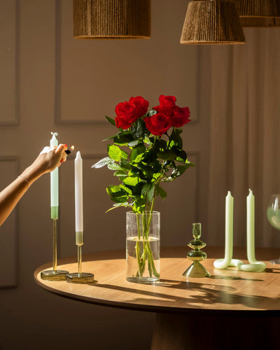 Eine Hand zündet eine weiße Kerze auf einem Holztisch an, der mit Roten Rosen (Inventar), grünen Kerzenhaltern und anderen Kerzen dekoriert ist, die alle sanft in warmem Licht leuchten.