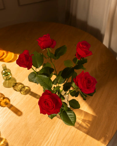 Ein Strauß Rote Rosen (Inventar) mit grünen Blättern liegt auf einem Holztisch im warmen Sonnenlicht und wirft weiche Schatten - eine zeitlose Liebeserklärung. Auf der linken Seite sind teilweise Glasflaschen und Orangenscheiben zu sehen.