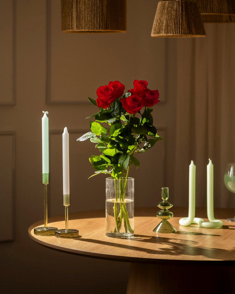 Ein Klassiker: Rote Rosen (Inventar) in klarer Glasvase auf rundem Holztisch, umgeben von grünen und weißen Kerzen in goldenen und gläsernen Haltern unter sanftem Licht - eine Liebeserklärung.