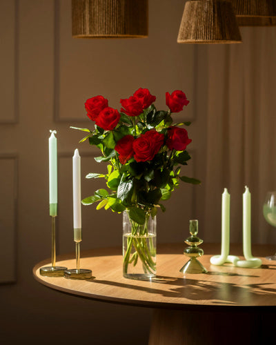 Eine klare Glasvase mit Roten Rosen (Inventar) steht auf einem runden Holztisch, umgeben von grünen und weißen Kerzen in verschiedenen Haltern bei warmer, weicher Beleuchtung - ein Klassiker, ideal für jede Liebeserklärung.