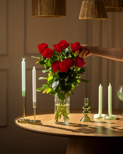 Eine Hand arrangiert "Rote Rosen (Inventar)" - ein echter Klassiker und eine perfekte Liebeserklärung - in einer klaren Glasvase auf einem runden Holztisch, mit grünen und weißen Kerzen in Kerzenhaltern, sanft beleuchtet von warmem Licht.