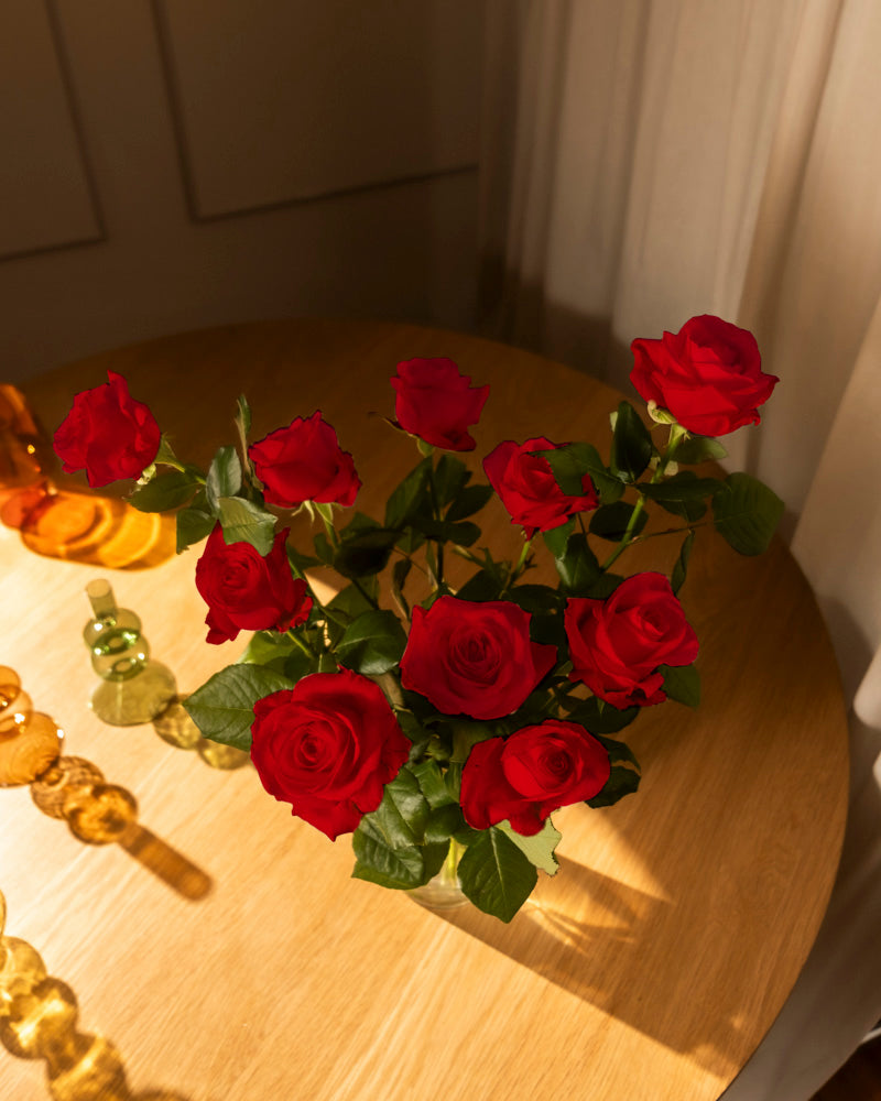 Ein Strauß Rote Rosen (Inventar) mit üppigen grünen Blättern steht in einer Glasvase auf einem hellen Holztisch, das Sonnenlicht fällt durch weiße Vorhänge. Bernsteinfarbenes Glasdekor sorgt für Wärme - ein zeitloser Klassiker als herzliche Liebeserklärung.