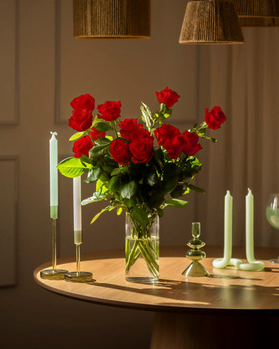 Ein Strauß Rote Rosen (Inventar) in einer Glasvase ziert einen Holztisch, umringt von hellgrünen Kerzen in dekorativen Haltern - eine zeitlose Liebeserklärung, sanft beleuchtet von warmem Licht und geflochtenen Lampen darüber.