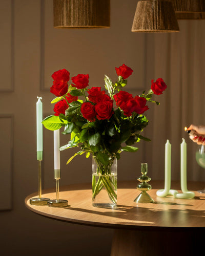 Ein Strauß Rote Rosen (Inventar) in einer Glasvase steht auf einem runden Holztisch neben grünen Kerzenhaltern; eine Hand zündet eine Kerze an und warmes Licht verbreitet gemütliche Atmosphäre im Raum.