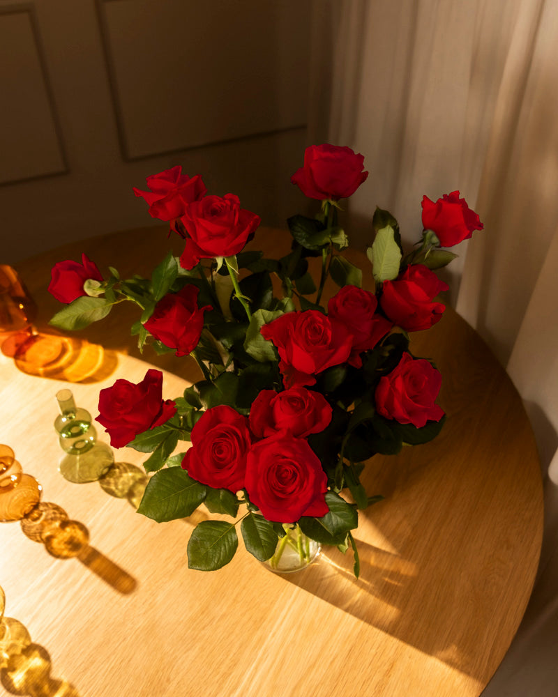 Ein Strauß Rote Rosen (Inventar) mit grünen Blättern in einer Glasvase steht auf einem runden Holztisch, der in warmes Sonnenlicht getaucht ist. Bernsteinfarbene und grüne Glasflaschen schmücken die Szene - ein echter Klassiker und eine stille Liebeserklärung.
