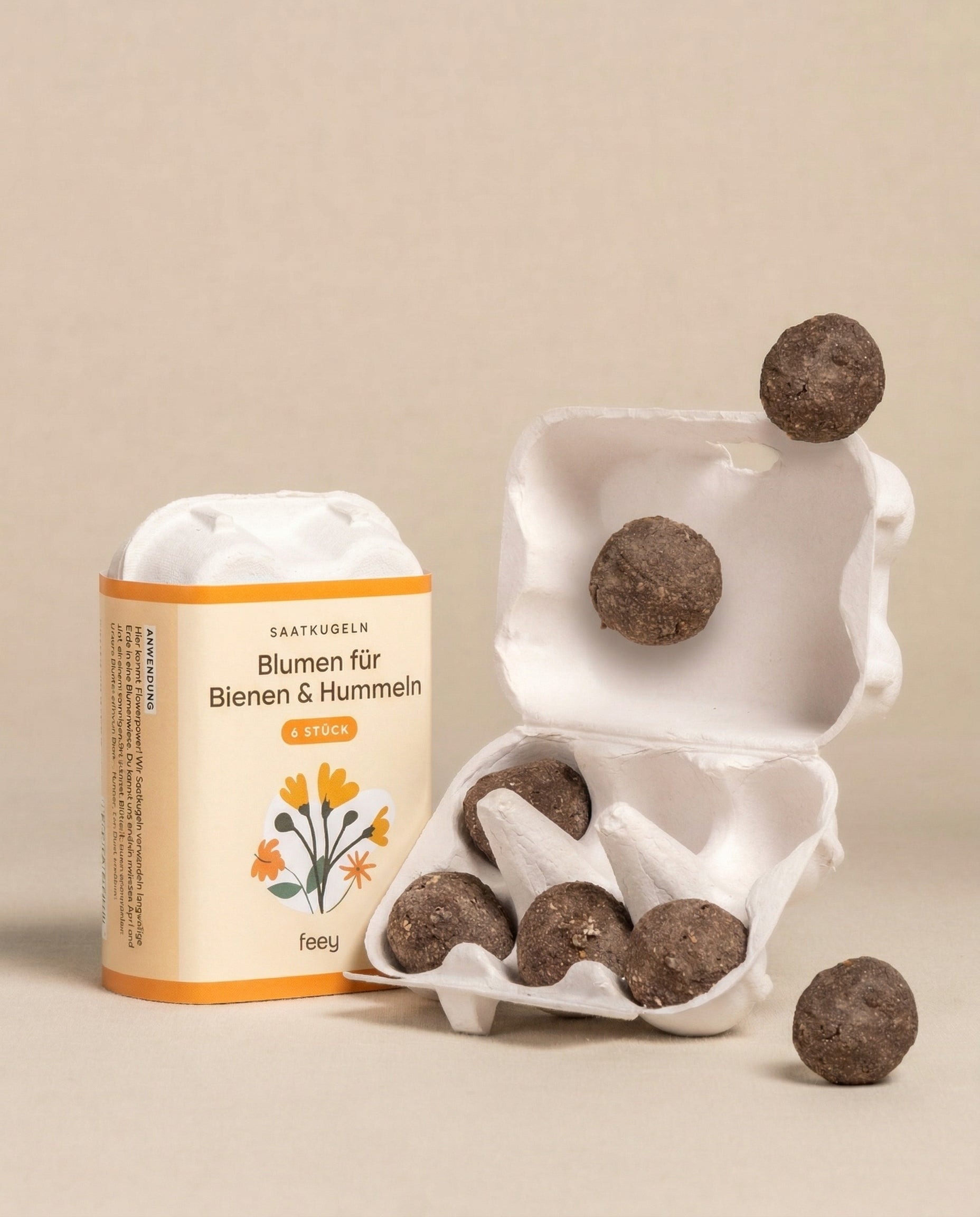 Ein kleiner offener Eierkarton mit runden braunen Saatkugeln für Feld- & Wiesenblumen steht neben einer beschrifteten Schachtel mit der Aufschrift 