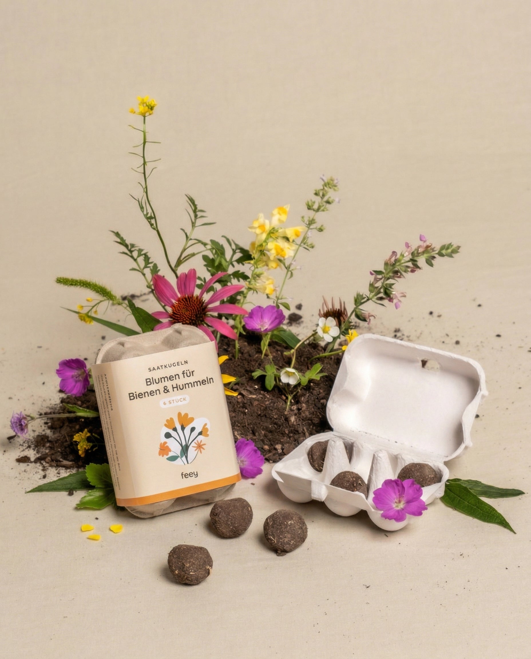 Eine Packung Saatkugeln für Feld- & Wiesenblumen und ein geöffneter Eierkarton mit Samenkugeln liegen auf dem Boden, umgeben von blühenden Wildblumen in lila, rosa und gelben Farbtönen, die im Hintergrund Bienen anlocken.