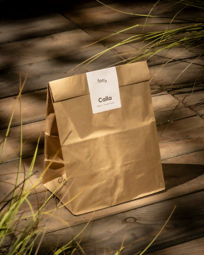Eine braune Papiertüte mit der Aufschrift „Calla (weiß)“ liegt auf einer Holzfläche, umgeben von langen grünen Zantedeschia-Blättern. Sonnenlicht wirft Schatten und schafft eine natürliche und ruhige Atmosphäre.
