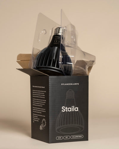 Eine schwarze Pflanzenlampe, die teilweise aus der geöffneten Schachtel genommen wurde, ist in durchsichtiges Plastik gehüllt. Die Verpackung trägt den Namen "Pflanzenlampe" und enthält in weißer Schrift Angaben zu gesunden Zimmerpflanzen.