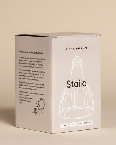 Eine beigefarbene Schachtel mit der Aufschrift "Pflanzenlampe" zeigt eine Strichzeichnung einer Lampe, Produktangaben (22W, E27, Vollspektrum) und einen deutschen Text, der erklärt, dass die Lampe Sonnenlicht für Zimmerpflanzen liefert.