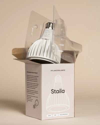 Eine Pflanzenlampe, die für Zimmerpflanzen bestimmt ist, wird teilweise aus einem "Staila"-Karton ausgepackt. Die Lampe verbleibt in ihrer durchsichtigen Kunststoffverpackung, und auf der Schachtel sind Produktinformationen angegeben. Ein großer Kühlkörper ist sichtbar.