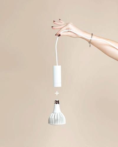 Eine Hand mit lackierten Nägeln hält eine weiße Pendelleuchte 'Corda' über einer weißen Glühbirne, mit einem Pluszeichen dazwischen, vor einem beigen Hintergrund.