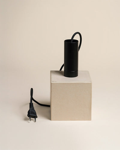 Eine schwarze zylindrische Pendelleuchte "Corda" mit Netzkabel und Stecker ruht auf einem beigen rechteckigen Kasten vor einem schlichten hellen Hintergrund.