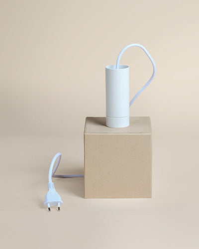 Eine weiße zylindrische Pendelleuchte "Corda" mit Netzkabel und Stecker befindet sich in einer schlichten braunen Schachtel auf beigem Hintergrund.