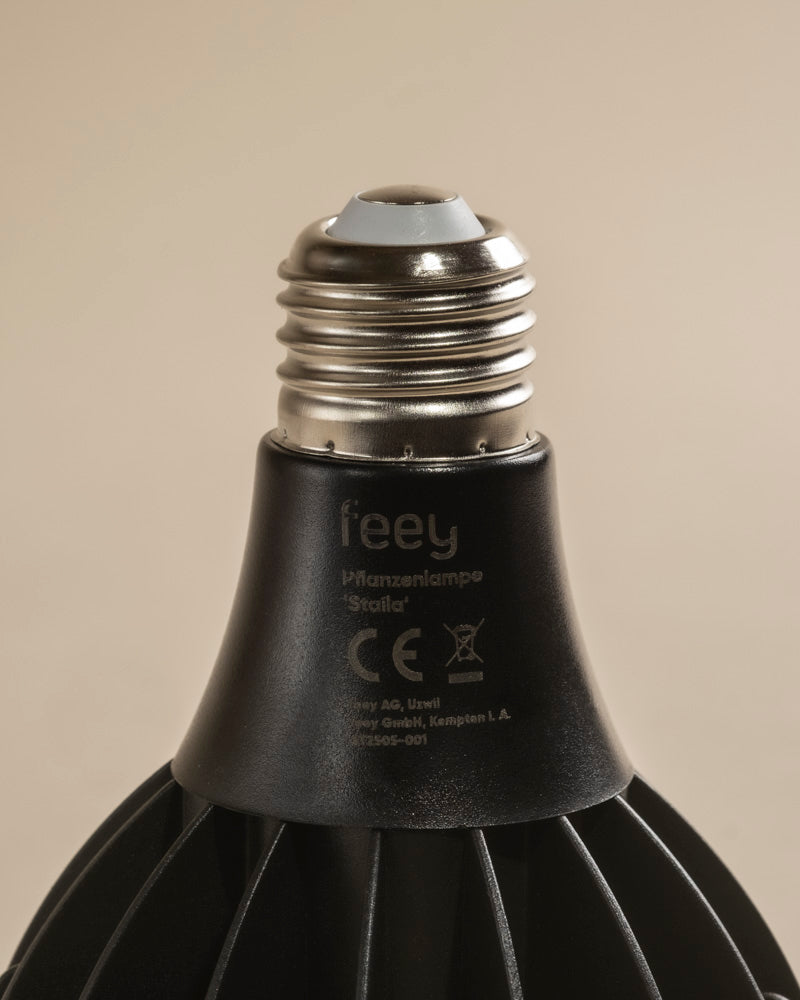 Nahaufnahme einer schwarzen LED-Pflanzenlampe mit metallischem Schraubsockel, die Produktdetails und die Zertifizierung auf dem Hals zeigt. Sie wurde entwickelt, um das Sonnenlicht für ein gesundes Wachstum von Zimmerpflanzen zu imitieren, und steht vor einem neutralen beigen Hintergrund.