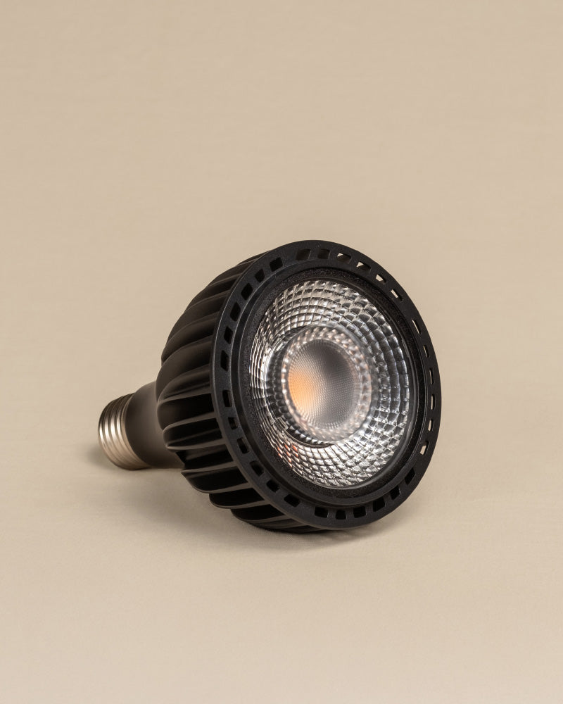 Eine schwarze Pflanzenlampe LED-Strahlerbirne mit metallischem Schraubsockel liegt auf der Seite vor einem schlichten beigen Hintergrund. Entwickelt, um optimales Sonnenlicht für blühende Zimmerpflanzen zu bieten, sind die Linse und die inneren Komponenten deutlich sichtbar.