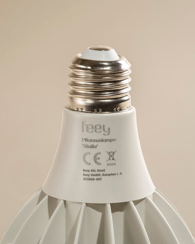 Nahaufnahme des Sockels einer weißen Pflanzenlampe mit einer Metallverschraubung und einem Etikett mit Modellangaben und Zertifizierungssymbolen. Sie ist ideal für Zimmerpflanzen und bietet sonnenlichtähnliches Licht. Vor einem neutralen beigen Hintergrund.
