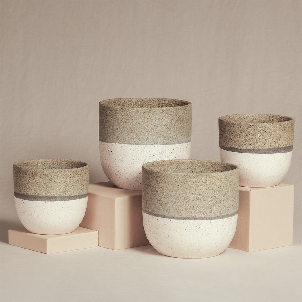 Ein Set aus vier handgefertigten, minimalistischen, runden Keramik-Pflanzgefäßen in verschiedenen Größen. Jedes Keramik-Topfset „Variado“ (2 × 18, 2 × 14) hat einen gesprenkelten, cremefarbenen Sockel mit einem hellbeigen Band an der Oberseite. Sie stehen auf hellrosa Sockeln vor einem beigen Hintergrund und eignen sich perfekt zur Präsentation Ihrer Zimmerpflanzen.