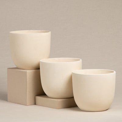 Drei minimalistische, cremefarbene Keramik-Topfsets „Direito“ (3 × 14) in verschiedenen Größen werden vor einem neutralen beigen Hintergrund präsentiert. Die handgefertigten Töpfe werden auf und neben zwei rechteckigen, beigen Blöcken platziert und bilden ein einfaches, elegantes Arrangement, das sich perfekt für Zimmerpflanzen eignet.