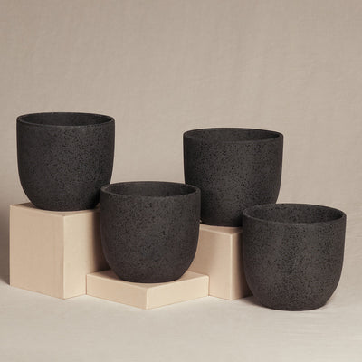 Auf und um drei beige Würfel unterschiedlicher Höhe herum sind vor einem neutralen Hintergrund vier schwarze Keramik-Topfsets „Direito“ (4 × 14) ausgestellt. Diese handgefertigten Töpfe zeichnen sich durch eine glatte, gesprenkelte Textur und ein minimalistisches Design aus, wodurch sie sich perfekt für die Präsentation von Zimmerpflanzen in jedem modernen Raum eignen.