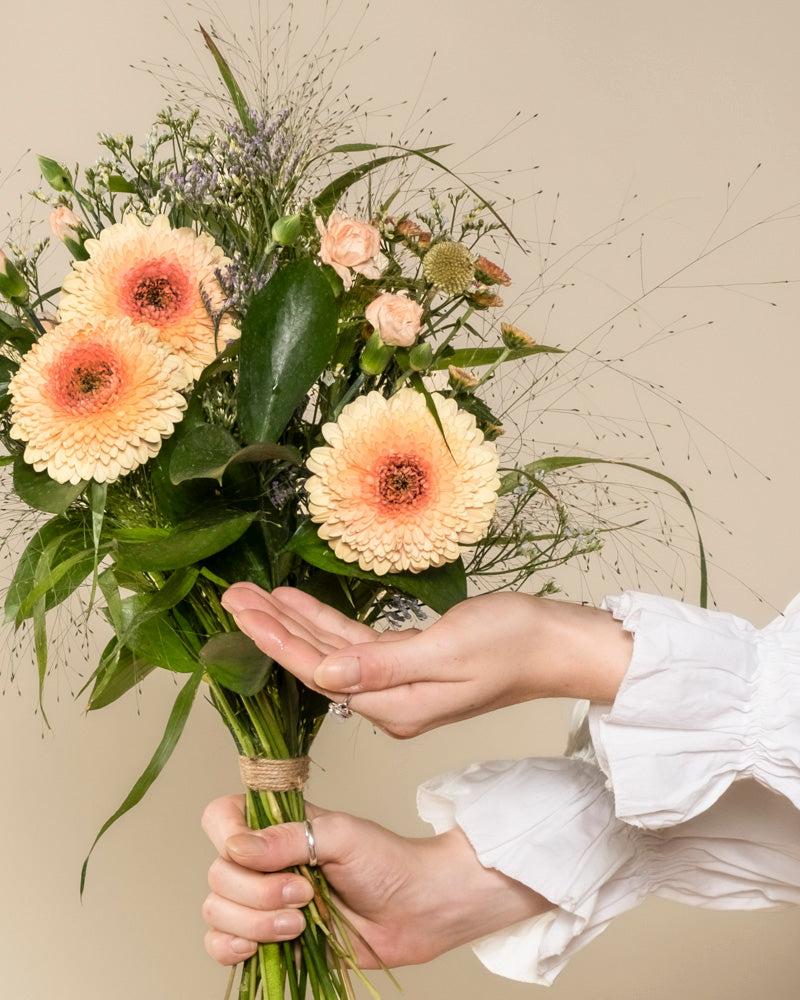 Eine Person mit weißer Rüschenärmel hält den "Überraschungs-Blumenstrauß" - einen liebevoll arrangierten Strauß aus hellen Gerbera, kleinen Rosen, Grün und Gräsern - vor neutralem Hintergrund in der Hand.