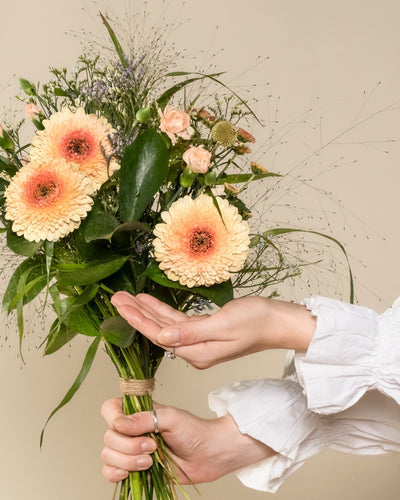 Eine Person mit weißer Rüschenärmel hält den "Überraschungs-Blumenstrauß" - einen liebevoll arrangierten Strauß aus hellen Gerbera, kleinen Rosen, Grün und Gräsern - vor neutralem Hintergrund in der Hand.